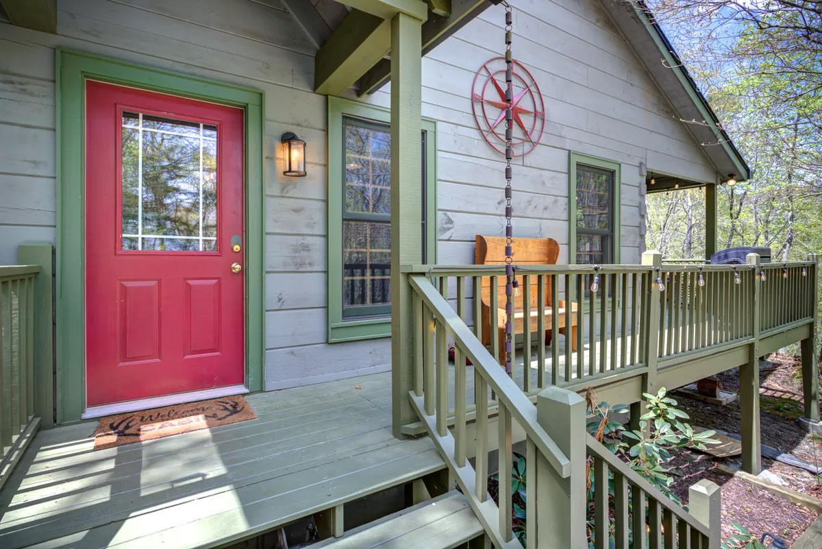 This image features the exterior of the cabin, highlighting the inviting red door, green accents, a decorative wheel, and a cozy swing bench on the porch. The surrounding greenery gives a strong sense of place and outdoor potential.