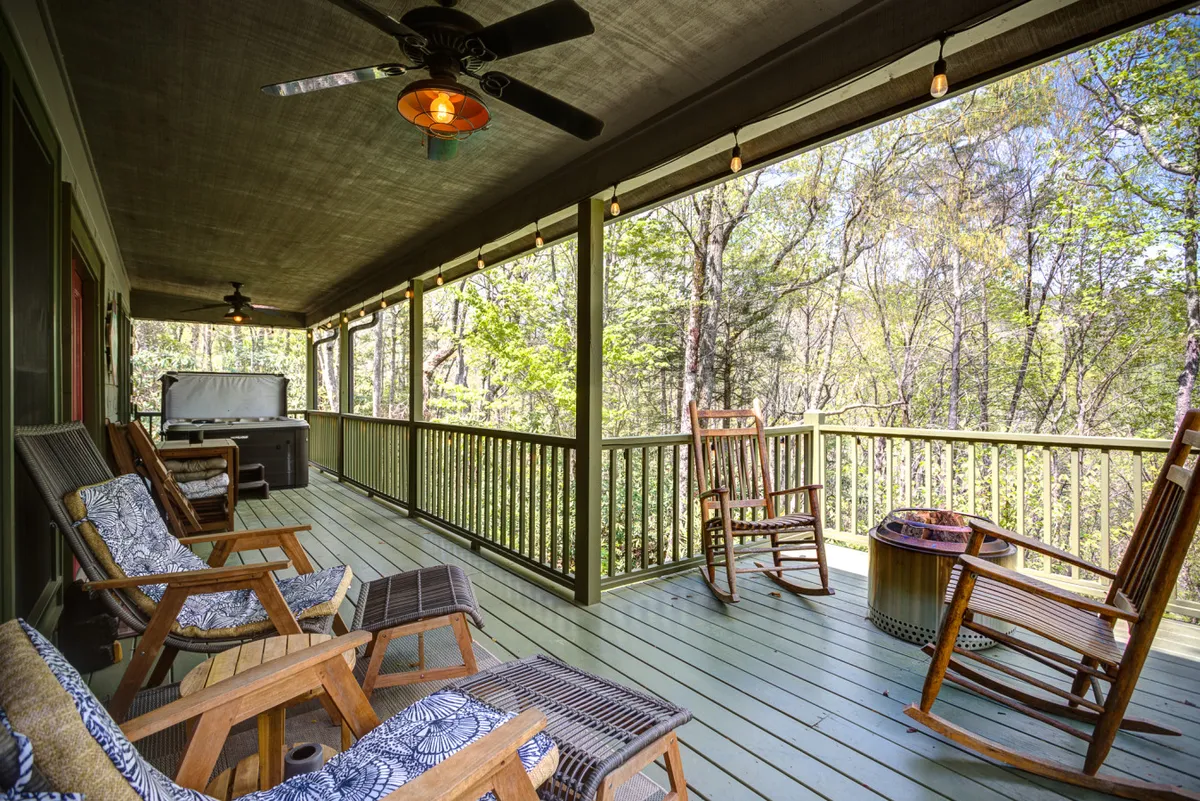 The image shows a spacious outdoor seating area with rocking chairs and wooden railing, surrounded by lush greenery. The space invites relaxation, socializing, and enjoying nature’s beauty.