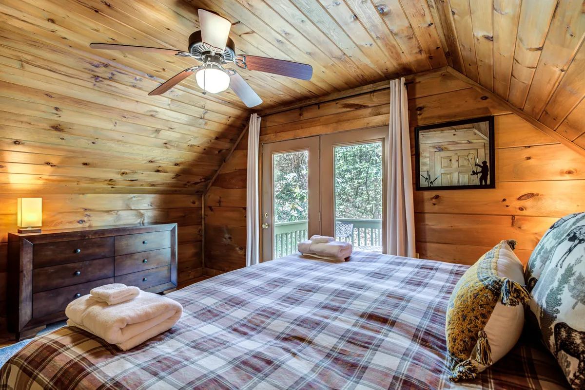 This image showcases a warmly decorated interior bedroom with wooden paneling, a plaid bedspread, and double doors opening out to a balcony. The space feels inviting and directly connects to the outdoors, hinting at the easy access to nature and nearby trails from within the cabin. The ceiling fan and cozy furnishings suggest comfort after a day exploring with pets.