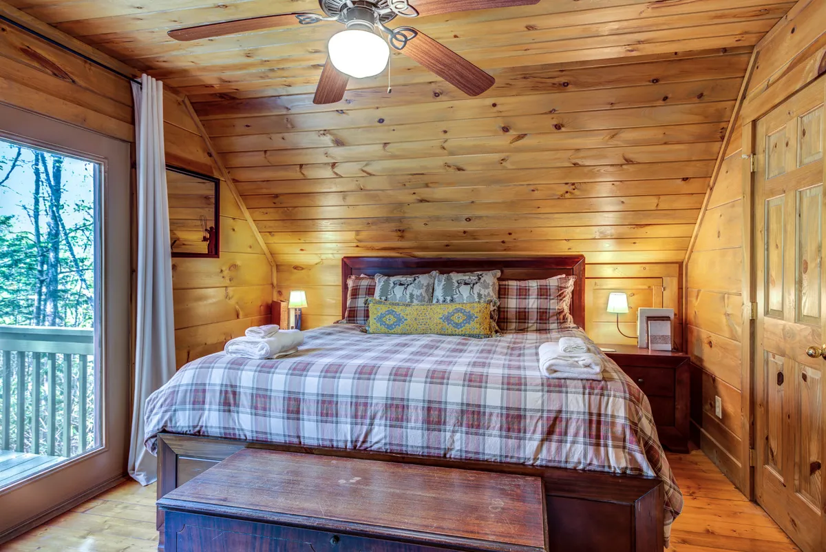 A cozy bedroom scene featuring a bed, wooden walls, a large window letting in abundant natural light, and a ceiling fan. Outside the window, trees and greenery are visible, emphasizing the cabin’s wooded, natural setting. The image evokes relaxation and a sense of being immersed in nature.