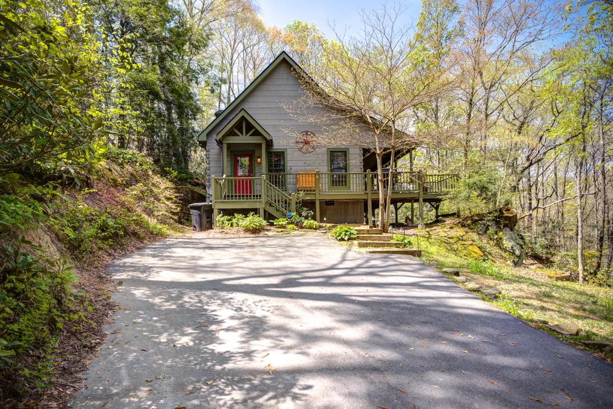 The image shows a charming log cabin surrounded by lush forest in Banner Elk, NC. A spacious front porch invites relaxation, with large windows offering a glimpse of the cozy interior. The setting is tranquil, bathed in soft natural light, and perfect for enjoying sunsets, outdoor gatherings, and the natural beauty of the Appalachian Mountains.