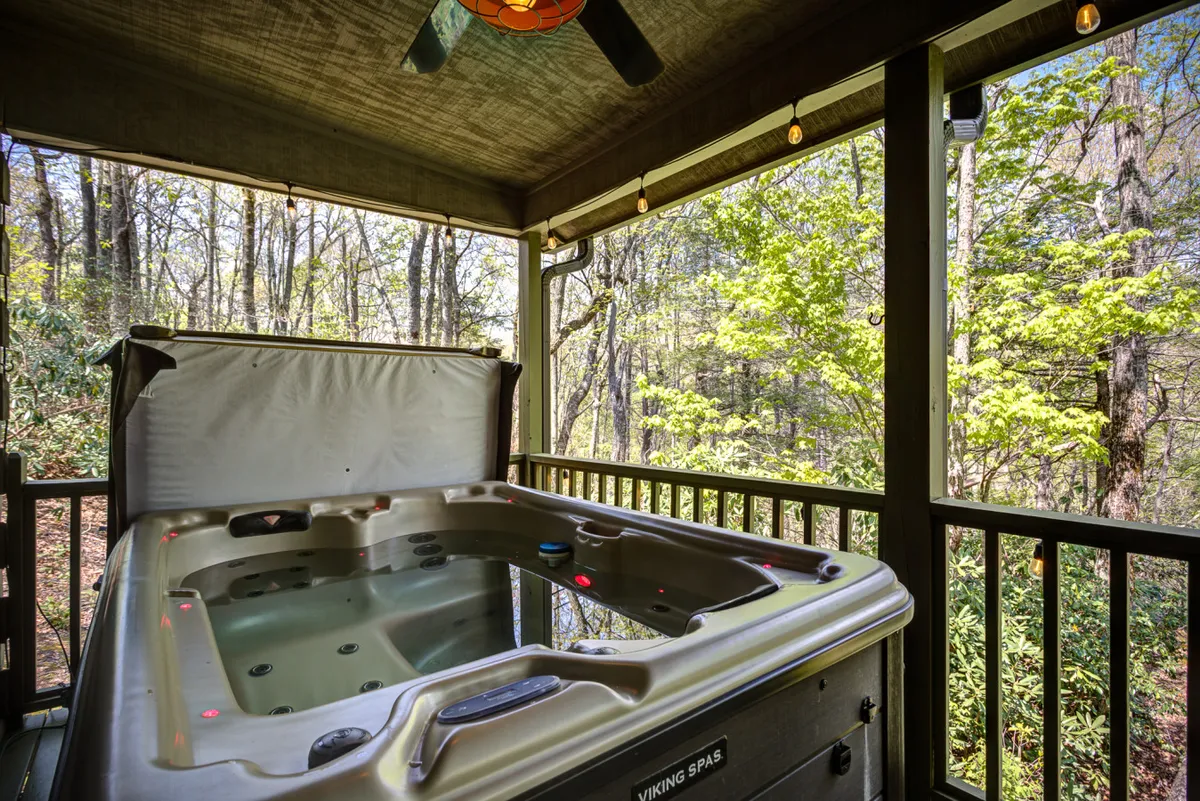 This image showcases a serene exterior deck with a two-person hot tub surrounded by lush greenery. Decorative lighting and a ceiling fan add a cozy touch, creating an inviting space to relax and take in the natural surroundings.