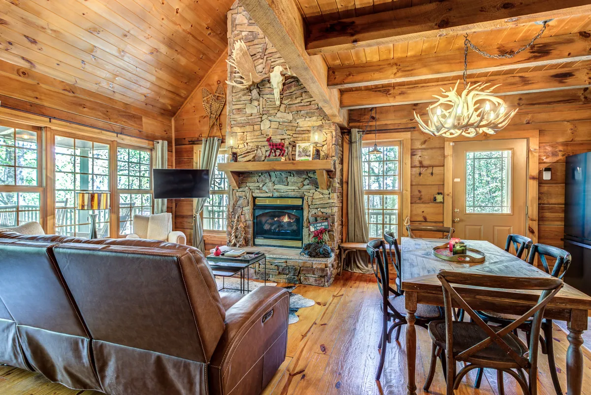 A spacious, light-filled interior living space with a stone fireplace, a large wooden dining table, and expansive windows looking out to the natural surroundings. The scene radiates warmth and communal comfort, ideal for shared meals or cozy evenings after outdoor activities.