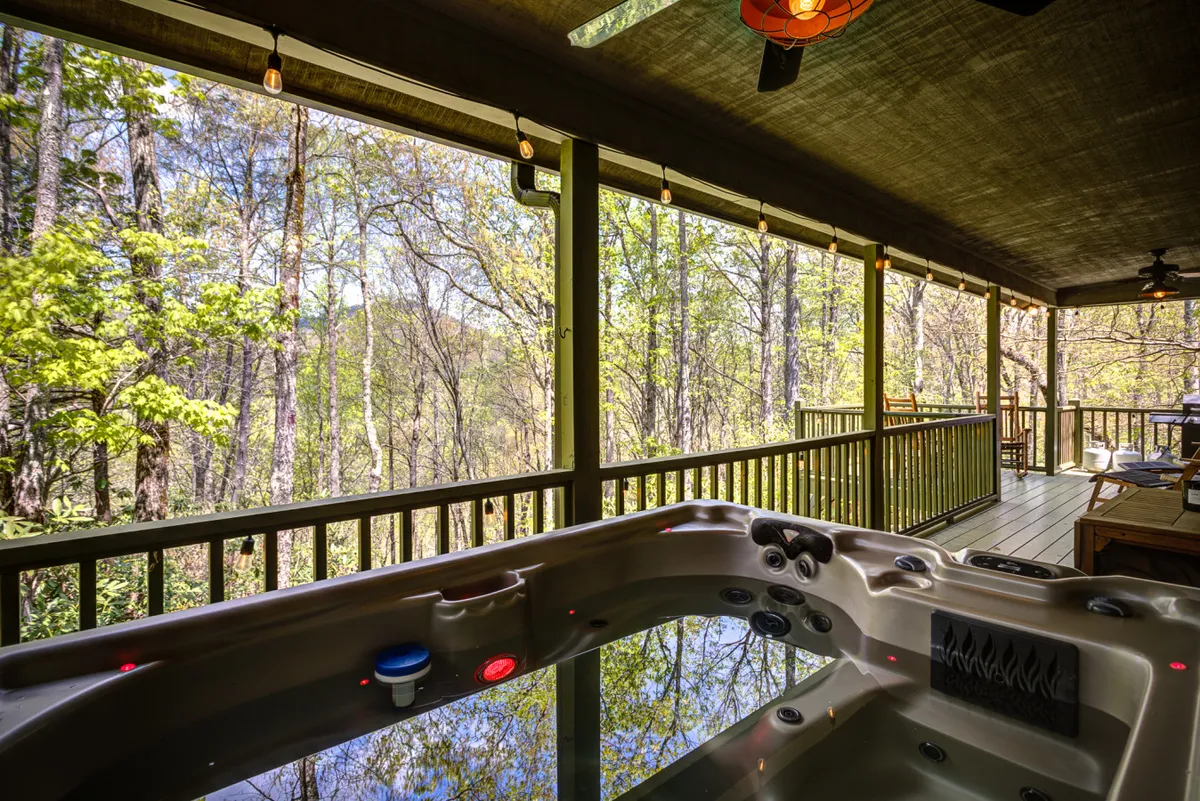 A tranquil exterior scene featuring a private hot tub on a wooden deck, surrounded by lush greenery and overlooking the serene mountain landscape. The hot tub is positioned for sunset views, with outdoor furniture inviting couples to relax together and enjoy the natural beauty.