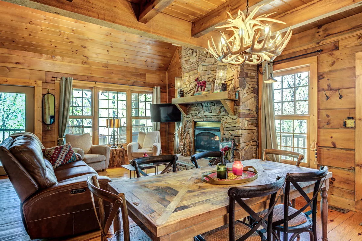 A warm and inviting living space featuring a stone fireplace, a rustic wooden dining table, an antler chandelier, and a comfortable seating area. The layout promotes easy flow between relaxation and dining, making it ideal for families and friends to gather, play games, or share meals.