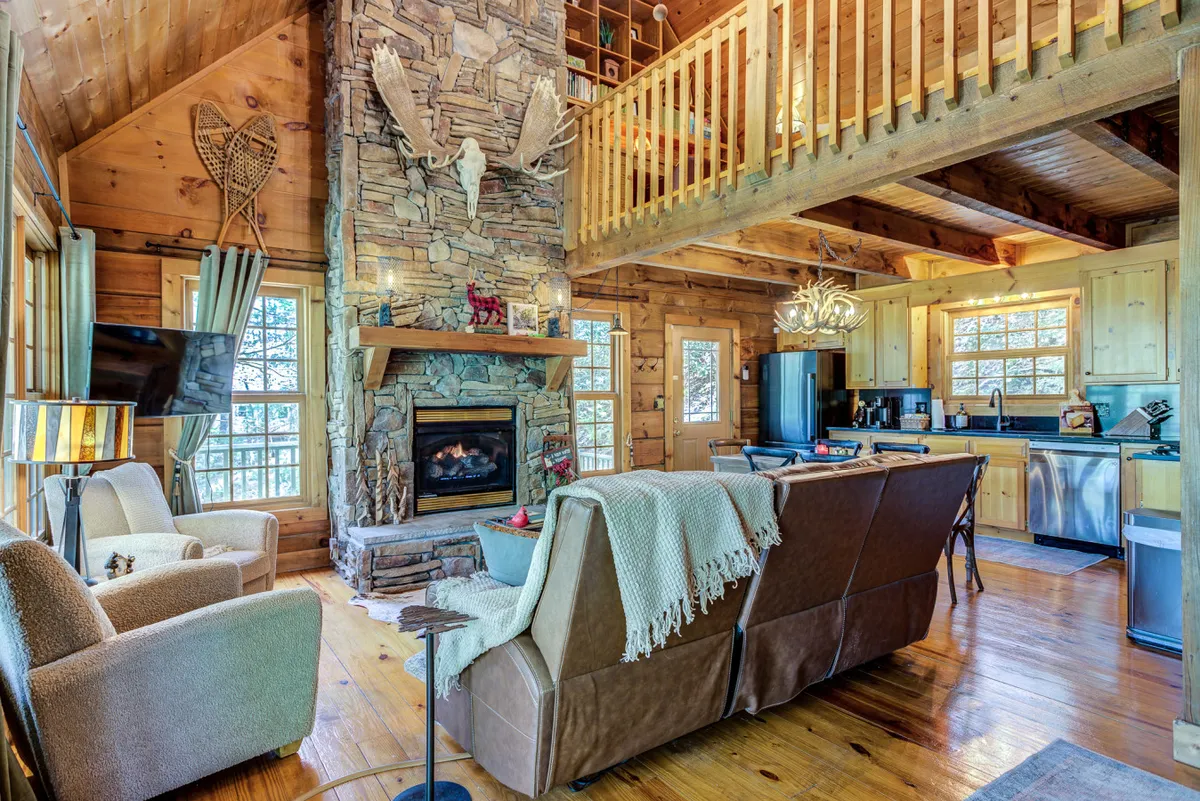 A spacious open-concept living area with a prominent stone fireplace as the centerpiece. The room features comfortable seating arranged to encourage conversation and relaxation. Abundant natural light pours in from large windows, highlighting the wooden beams and rustic decor. The space flows seamlessly into the kitchen area, emphasizing togetherness and connection.