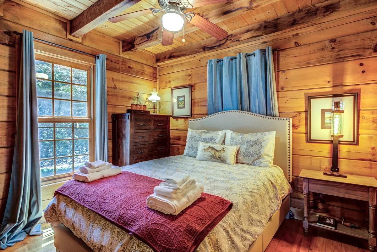 A queen bedroom with a comfortable bed topped with soft linens, positioned beside a large window. The room is decorated with a dresser, a lamp, and framed artwork, and is bathed in natural light, suggesting a peaceful, restful space ideal for children, friends, or guests.