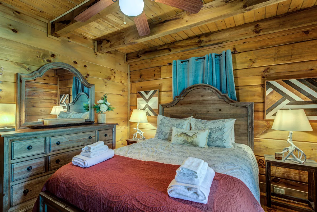 A warmly lit bedroom featuring a king-sized wooden bed frame adorned with plush decorative pillows and soft blankets. A dressing table with a mirror sits nearby, alongside tasteful artwork and softly glowing lamps. Sunlight filters in, highlighting the cozy, inviting atmosphere and the rustic decor touches that evoke both comfort and mountain charm.
