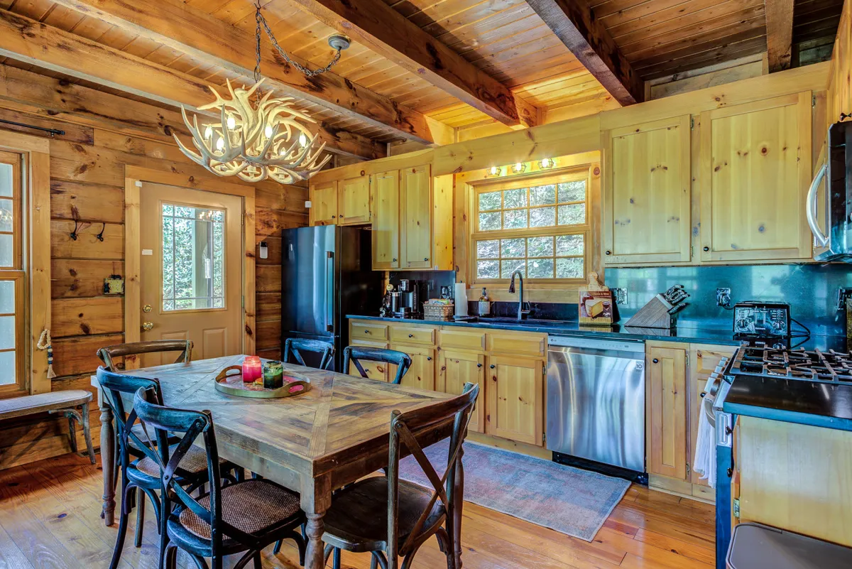 The space features a cozy wooden dining table beneath an antler chandelier, flanked by a window that brings in natural light and classic kitchen cabinetry for a welcoming, homey feel. The set-up suggests communal dining and the versatility of the space for various occasions.