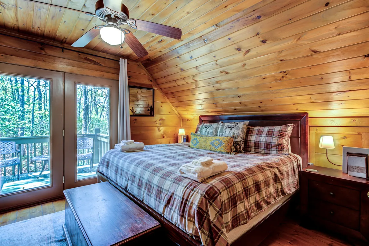 A tranquil bedroom with a wooden bed frame, decorative pillows, and wood-paneled walls. Large windows offer abundant natural light and views of the outdoors, while a ceiling fan and cozy blankets provide comfort for all seasons. The mood is peaceful and homely, ideal for relaxation and morning routines.