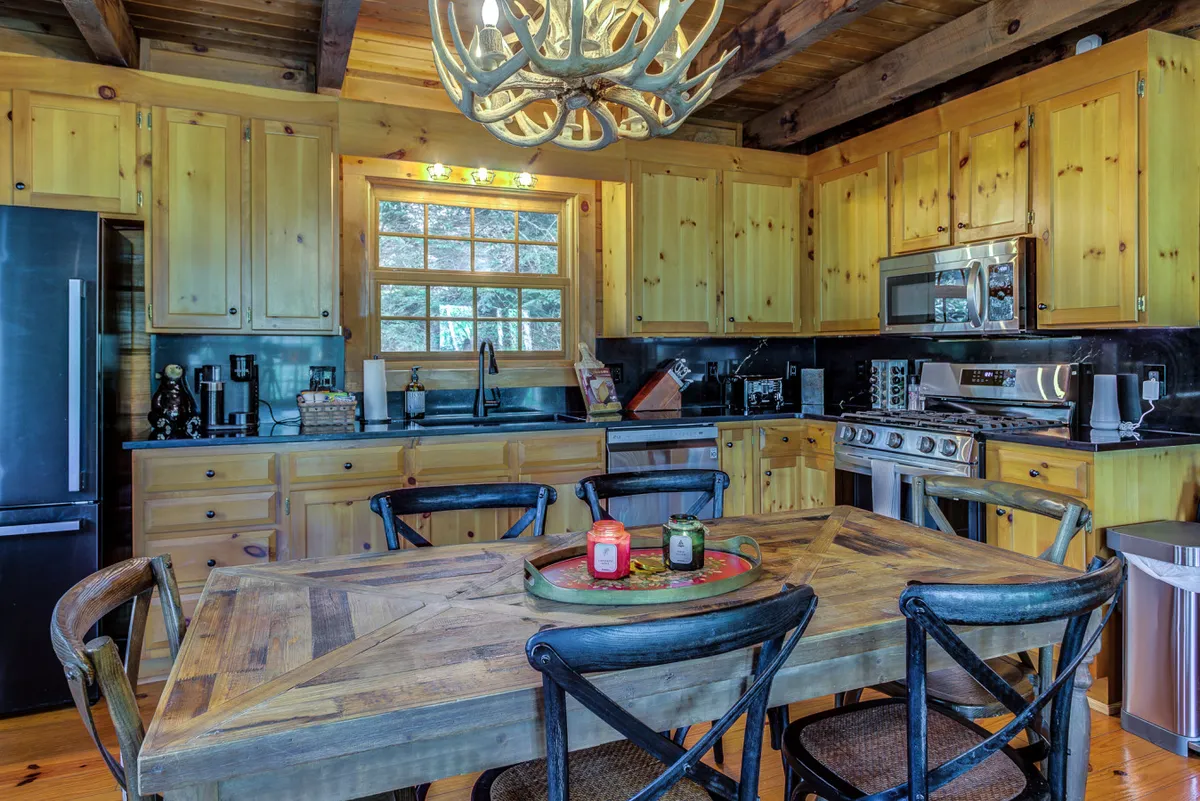 This image showcases a rustic wooden dining table set beneath an antler chandelier, with black appliances and wooden cabinetry visible in the background. Natural light pours in through a large window, illuminating the space and emphasizing the kitchen and dining area as an inviting hub for gathering.